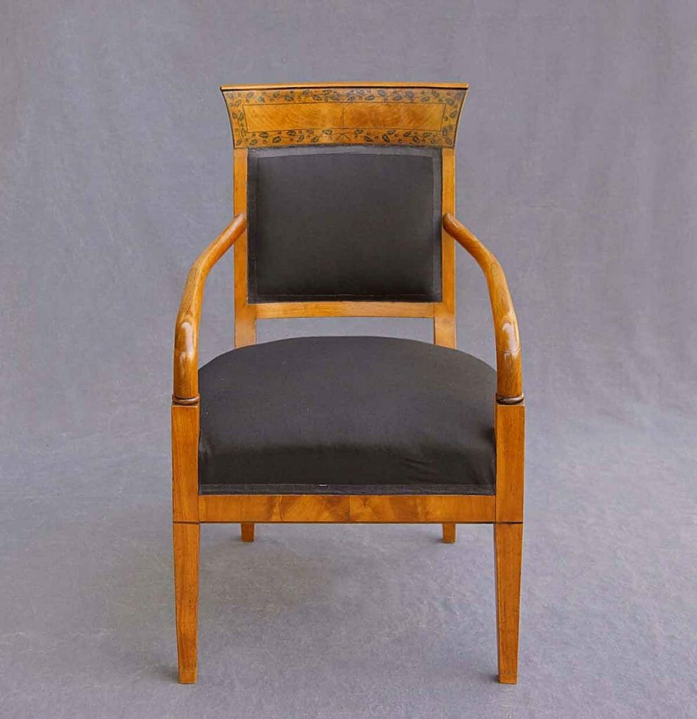 Auf dem Bild ist ein Armlehn-Sessel von vorne zu sehen. Er ist schwarz bezogen und aus Kirschbaum. Die Rückenlehne ist mit Schwarzlotmalerei bemalt.