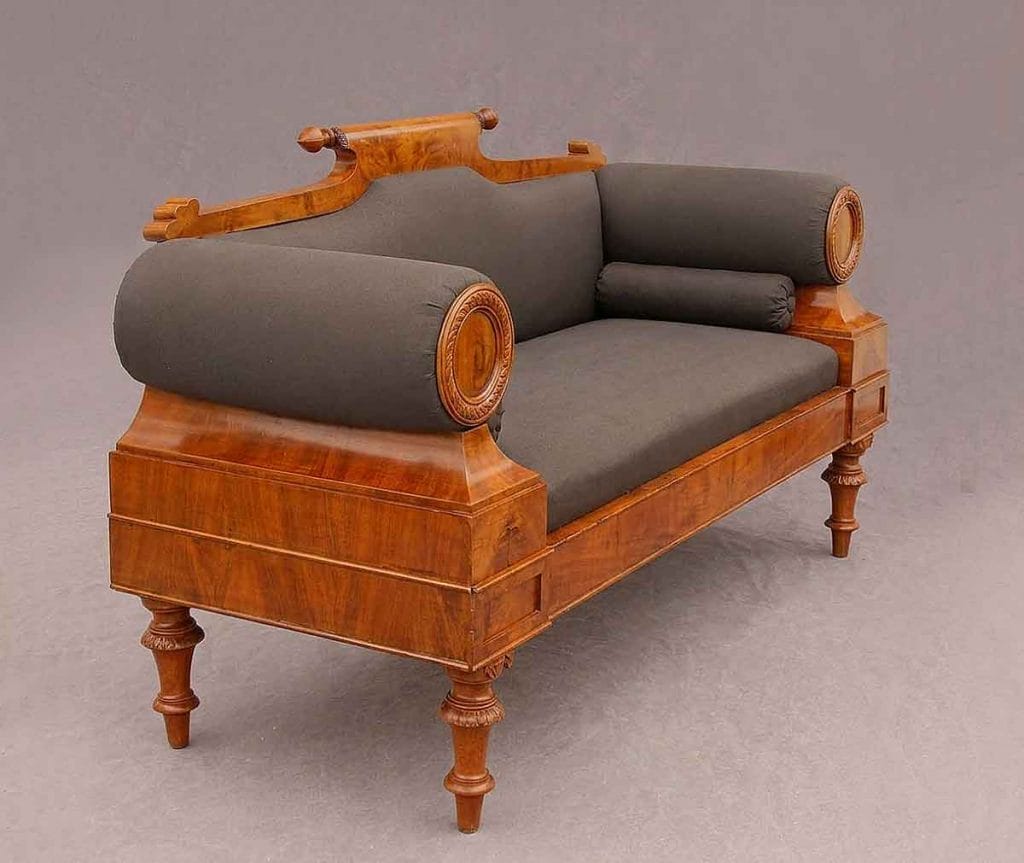 Biedermeier Sofa Nordeutsch4 Perspektivische Außenansicht eines antiken Biedermeier-Sofas mit poliertem Holzrahmen und dunkelgrauer Vorpolsterung, einschließlich zweier Rollkissen.