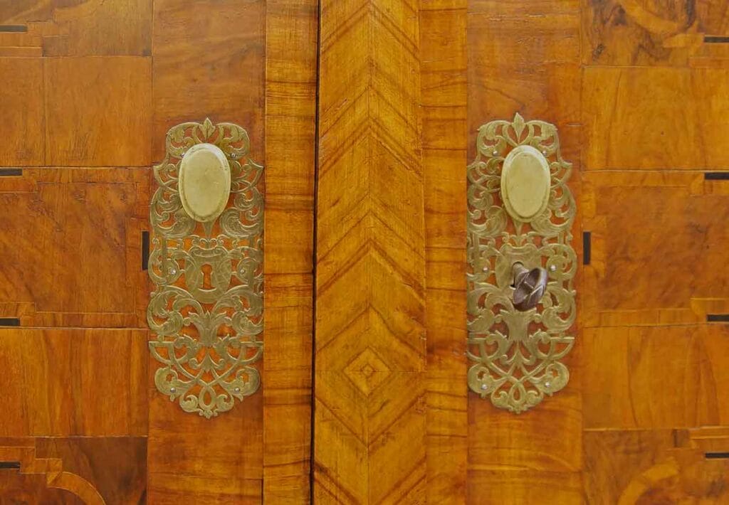 Barock-Schrank3 Nahaufnahme von zwei verzierten Messingbeschlägen und Schlüsselschildern an einer furnierten Holzschranktür im Barockstil.