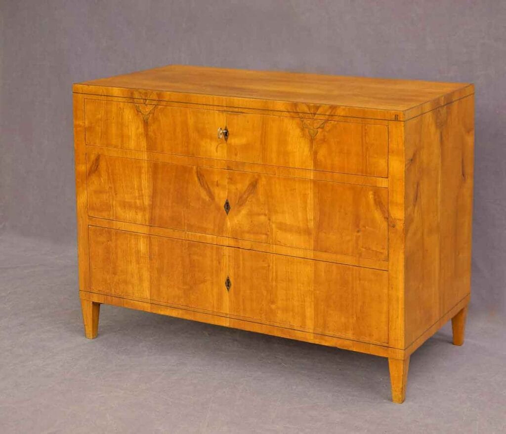 Biedermeier-Kommode-Kirsch Biedermeier-Kommode aus Kirschbaumholz in Dreiviertelansicht mit drei Schubladen und steckendem Schlüssel, vor grauem Hintergrund