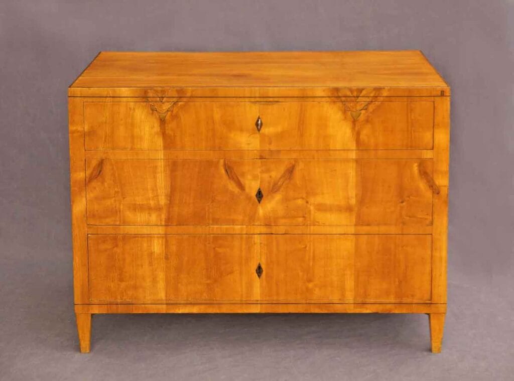 Biedermeier-Kommode-Kirsch1 Kommode aus Kirschbaumholz in Frontalansicht mit drei Schubladen und rautenförmigen Schlüsselschildern vor grauem Hintergrund