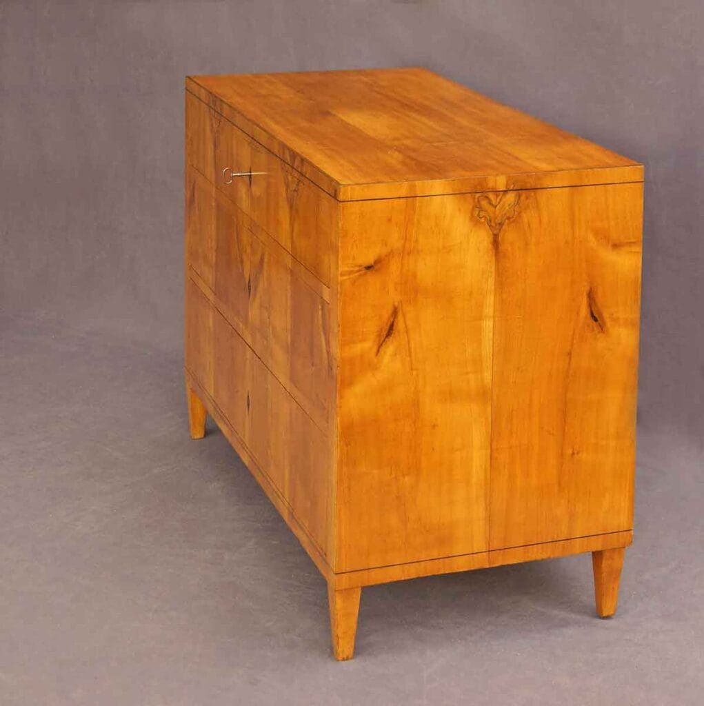 Biedermeier-Kommode-Kirsch2 Biedermeier-Kommode aus Kirschbaumholz in Seitenansicht von links mit drei Schubladen und steckendem Schlüssel, vor grauem Hintergrund.